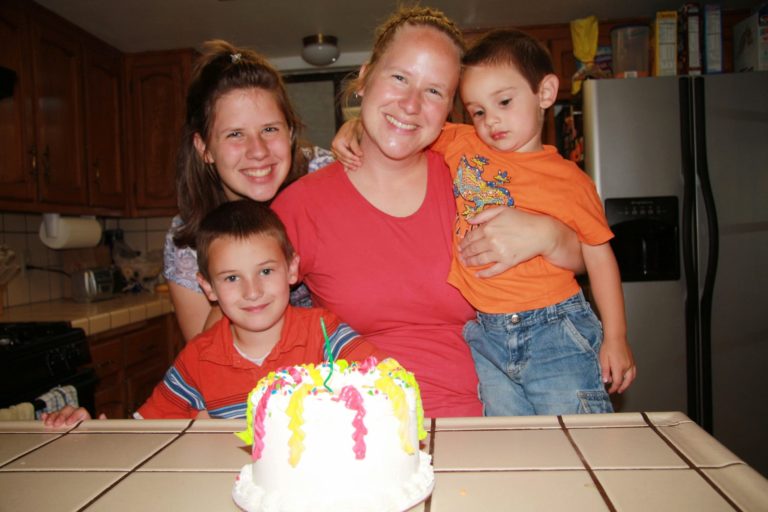 Me holding my two sons with my little sister looking over my shoulder as we stand behind my birthday cake.